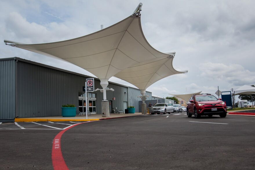 The entrance to the South Terminal at Austin-Bergstrom International Airport. A large white canopy would create shade, but this picture was taken on a cloudy day. Cars are parked in front. A car is pulling away.