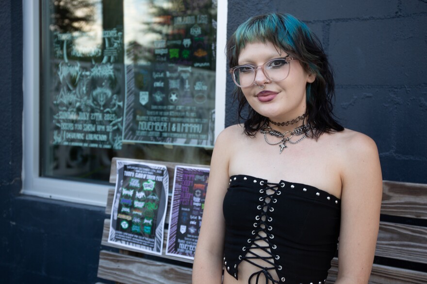 A person with shaggy black and blue hair and glasses sits on a bench outside a brewery. There are show posters pasted up behind them.