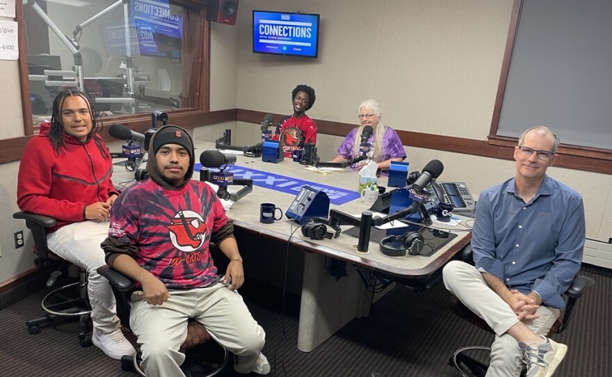 Five smiling people sit around a table in a radio talk studio: A young man front left has short black braids and is wearing a red hoodie, khaki pants, and white sneakers; a young man front center has a black mustache and is wearing a black hat with a red logo, a black hood, a red and black t-shirt with a red cat graphic, khaki pants, and white sneakers; a young man black left has short black hair and is wearing a red and black t-shirt with a red cat graphic; a woman back center has long white hair and is wearing glasses, a purple t-shirt with a white Rochester Flower City logo; a man at right has very short dark hair and is wearing a blue button-down shirt, white linen pants, and sneakers 