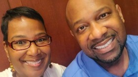 black woman with dark hair and glasses stands next to a bald black man, both smiling 