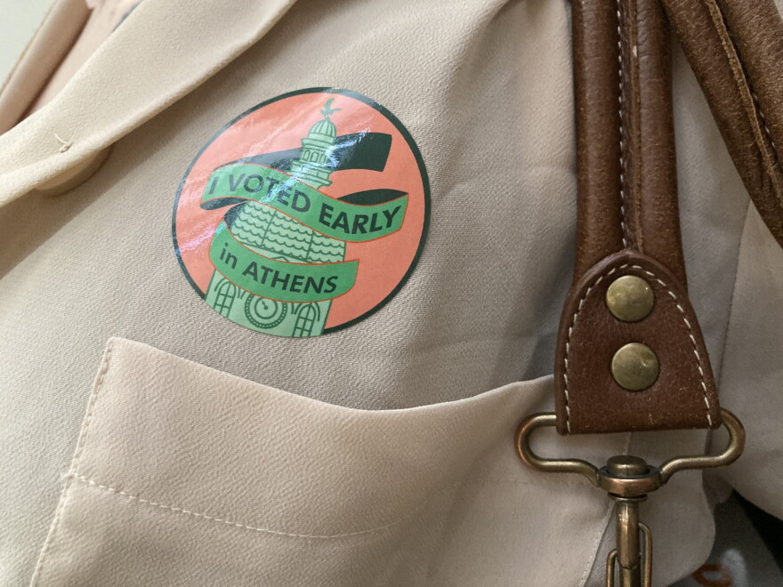 A close-up of an "I voted early in Athens" sticker on someone's beige blouse, next to the brown leather straps of their handbag. 