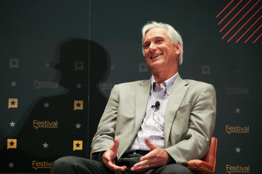 A man in a suit sits in a chair with a black screen backdrop in the middle of speaking.