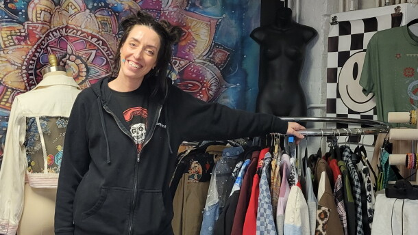 A woman in a black top and green checkered pants smiles next to a rack of clothes