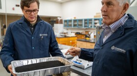 Two men in dark blue lab coats talk while gesturing to a pan full of a black coarse material.