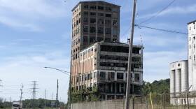 An abandoned, multi-story industrial building with broken windows stands behind a chain-link fence next to a highway.