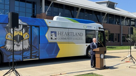 A man stands speaking at a podium, with a large bus behind him. The Connect Transit bus has a hawk on it and says Heartland Community College on the windows.