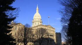 Wide view of the state capitol building. It is lit up by the sun, with blue sky behind, and the building is framed by trees in the foreground