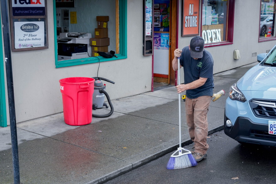 A man sweeps a sidewalk in front of a business missing its store-front window.