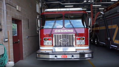 This is the front of a fire truck with the name of the town, Ossining, across the front. The fire truck is in a brick garage with another fire truck next to it. 