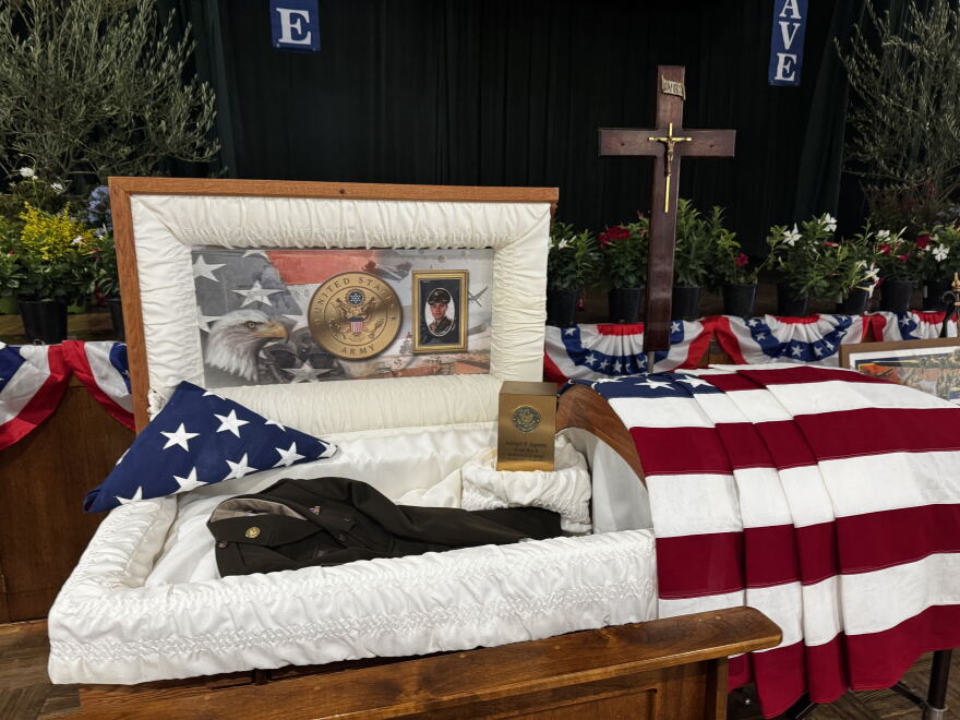 Casket with photos of Sgt. Adolph Aguirre and his uniform. American flag draped over. 