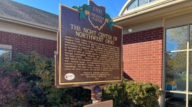 An Ohio Historical Marker stands outside a red brick building. A plaque with braille sits beneath the paragraph of text and a green audio kiosk stands beside it.