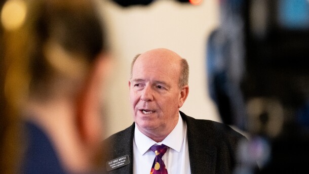 A balding man in a gray suit framed between a television camera and the camera operator.
