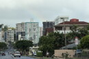 cars drive on a rainy road with apartments in the background and a faint rainbow in the cloudy sky