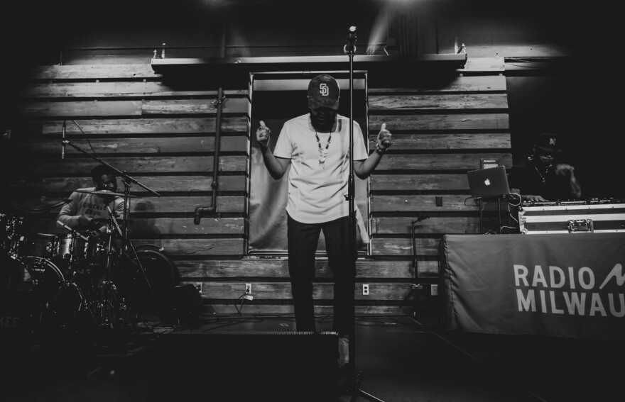  A man in a white t-shirt and baseball cap poses on stage with his head bowed and his hands extended to the crowd while a DJ operates turntables next to him.