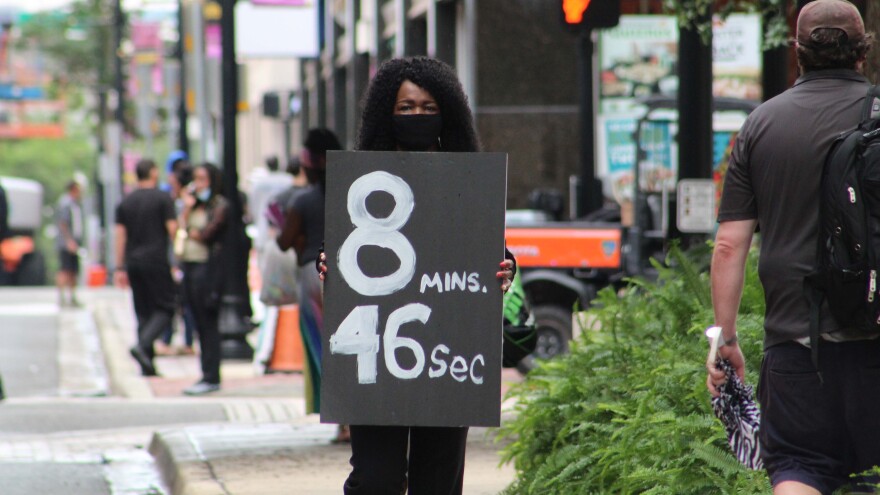 Woman wearing mask holds a sign with 8 minutes 46 seconds on it