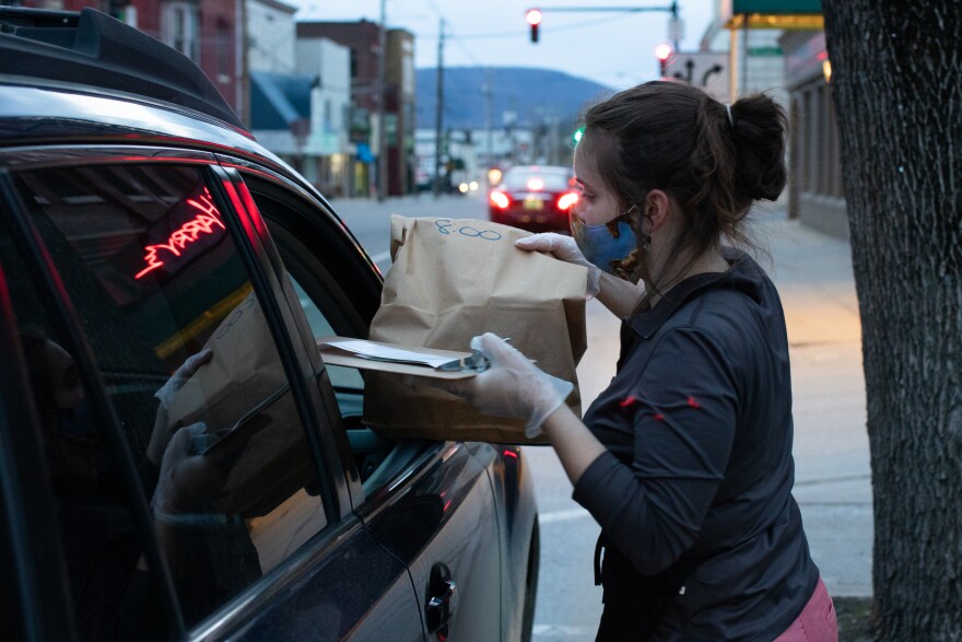 A person wearing a face mask delivering a paper bag to a car window.