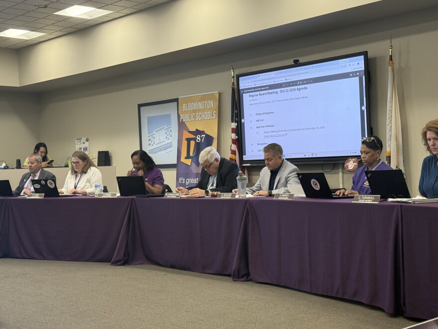 District 87 school board members and the superintendent sit at a long table draped in purple with laptops and nameplates, beneath a screen showing the Oct. 21 meeting agenda and beside a Bloomington Public Schools banner.