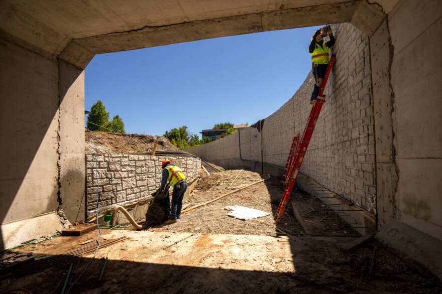 At the end of a tunnel, one construction worker bends toward the ground while another stands on a ladder to work. 