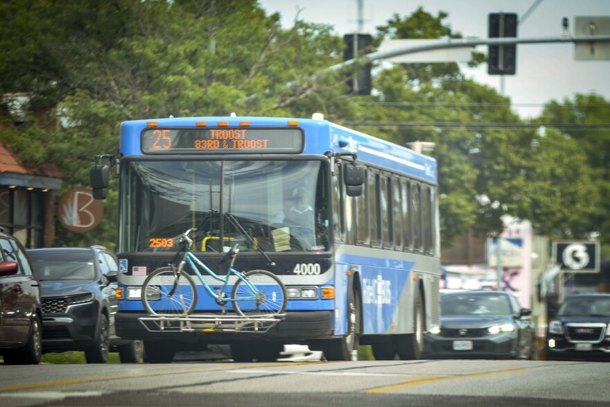 A blue transit bus moves along a busy street. On its sign designation, it reads "83rd & Troost. It appears to be moving past an intersection where other cars are just behind it. A bicycle sits on the front rack of the bus.