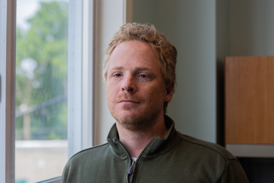 A person with short, light blond hair and light facial hair stands indoors near a window, looking slightly forward with a neutral, thoughtful expression. They are wearing a dark green quarter-zip sweater. Soft daylight from the window to their left lights one side of their face, while the background is softly blurred, showing a pale wall and part of a wooden cabinet. Greenery is visible outside the window.