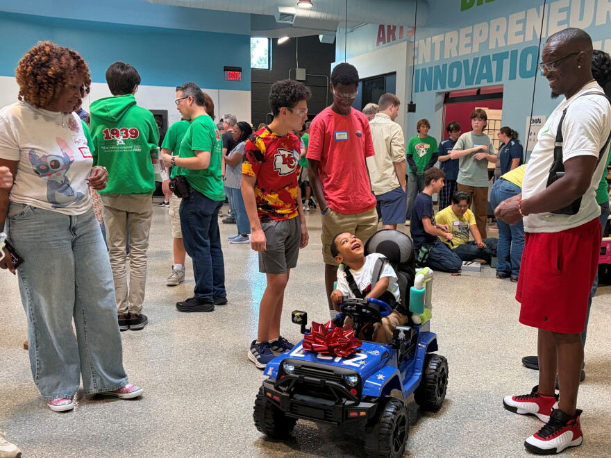 A woman stands looking at a child seated in a battery-operated toy car. The child is looking up and smiling at a man who is looking down and smiling at the child. Other people are standing an sitting in the background