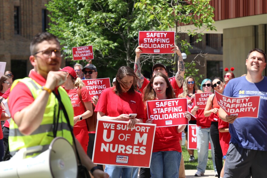 A group of people participate in an outdoor rally in support of University of Michigan nurses. In the foreground, a man in a neon safety vest speaks into a megaphone. Behind him, several individuals in red shirts hold up red and white signs that read "SAFETY STAFFING SOLIDARITY" and "WE SUPPORT U OF M NURSES." To the right, a man in a blue Michigan Medicine t-shirt holds a similar sign. The group is standing in a paved area with green trees and university buildings in the background.