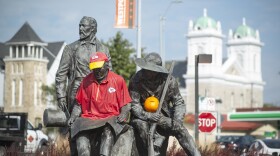 A bronze statue outside near a city street shows three men in pioneer garb shows one of the men wearing a modern Kansas City Chiefs football jersey and another character holding a pumpkin.