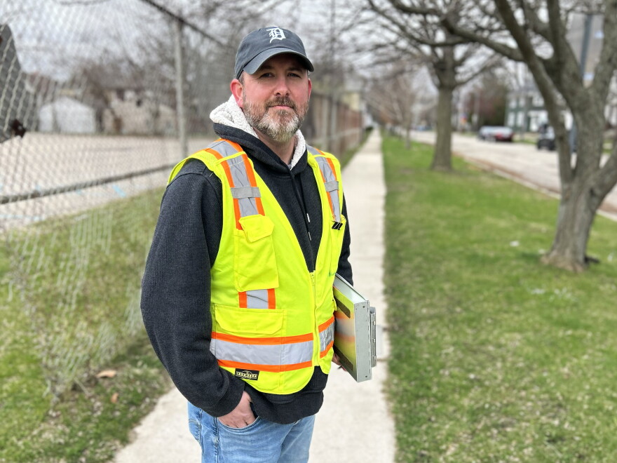 Man in a ballcap and greenish-yellow safety vest with clipboard under one arm smiles subtly while facing the camera