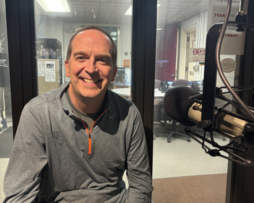 The top half of a man sitting in the Public Radio 90 studio, smiling next to a microphone