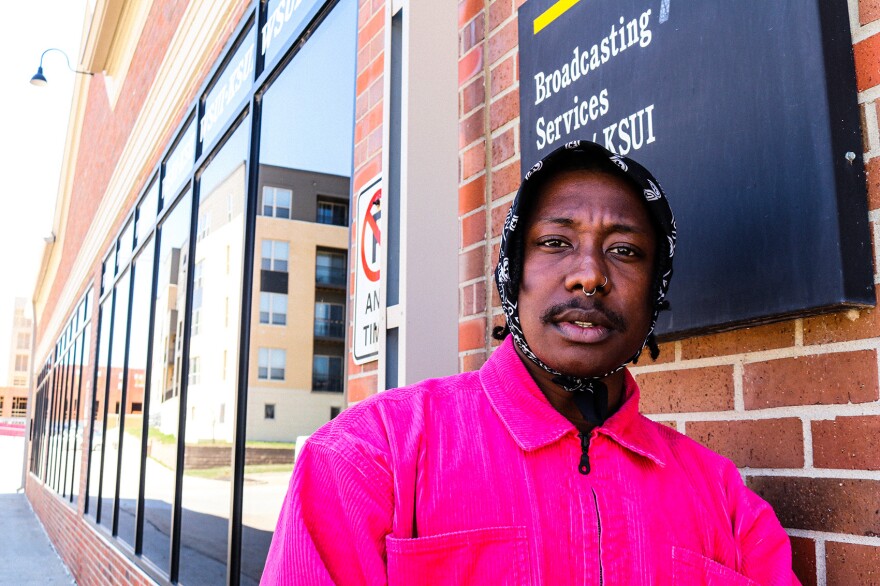 A man in a black bandanna and pink corduroy jacket stands outside of a radio station