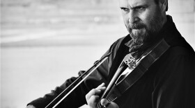 A man with a beard and a serious expression plays the violin in a black and white image.