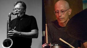 Two guys wearing black t-shirts. The one on the left playing a saxophone and the one on the right playing drums.