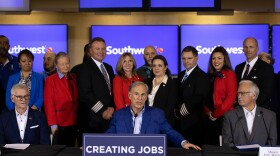 A man sits at a table speaking flanked by two other men on either side. Behind him is a line of people, some in airline captain uniforms, others in red jackets. A sign on the table reads 'creating jobs'