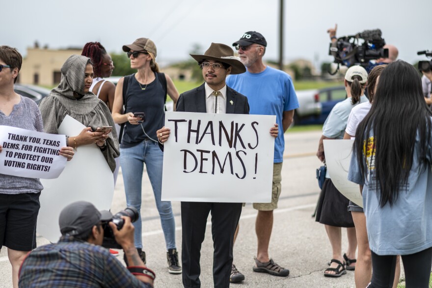 A man in a cowboy hat reads a sign reading "Thanks Dems!" as he stands in a small crowd of people with cameras and signs, including another that says "Thank you, Texas Dems, for fighting to protect our voting rights!"