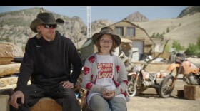 Dan and Lexy Meade sit on a wood block with a barn and several old motorcycles behind them. Dan has a reddish-blonde beard and sunglasses and wears a black hoodie and dark jeans. Lexy wears jeans, a wide-brimmed hat, and a shirt that says "lovers gonna love."