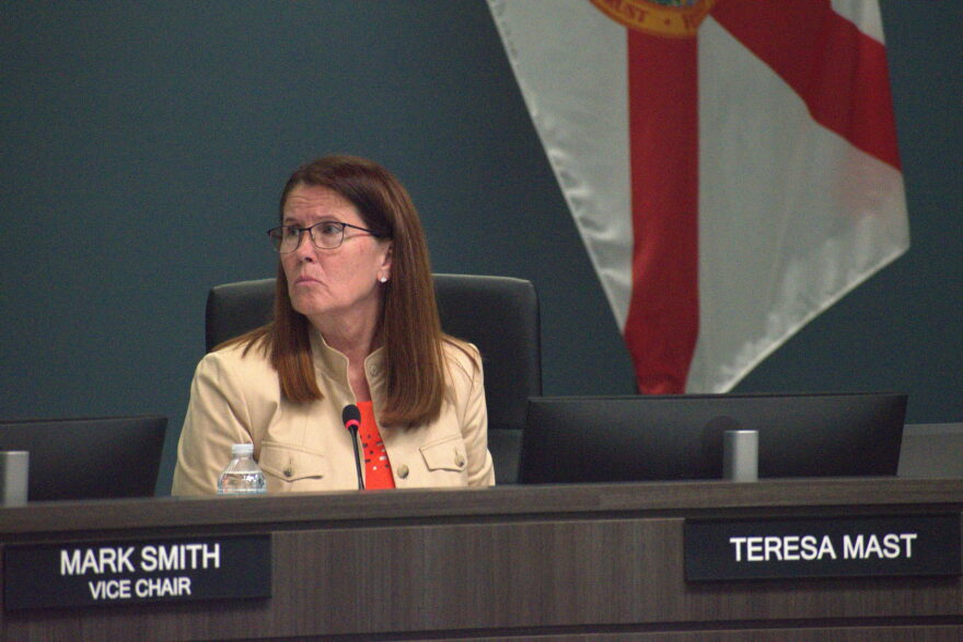 A woman in a bright orange shirt and tan jacket is sitting down at a brown desk in front of a black desktop computer and tiny microphone. 