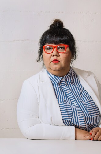 Virgie Tovar, with her hair tied into a bun, wears a white blazer and white and blue stripped button up.