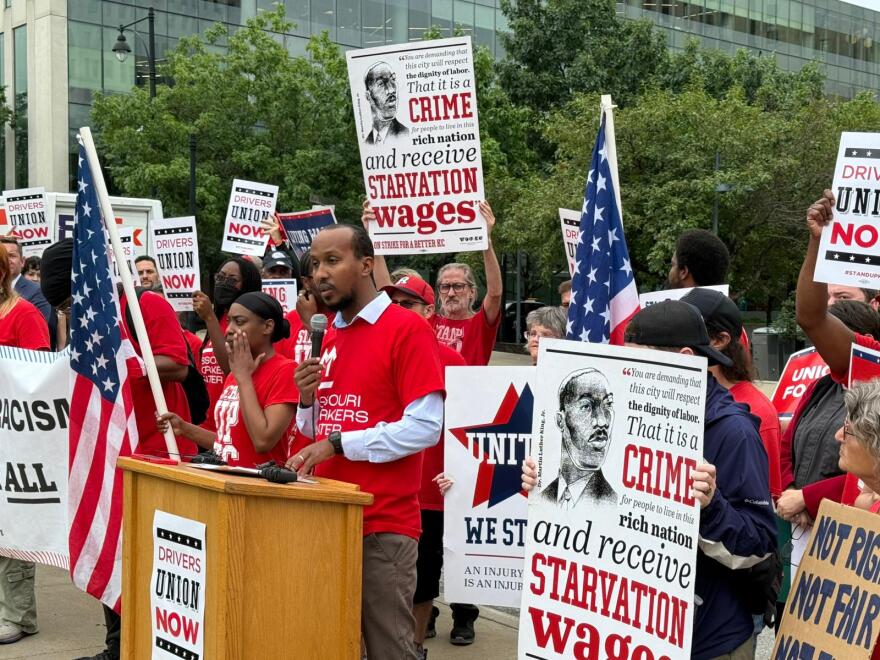 A man stands at a podium with a microphone in hand. Near the man, several others gather holding signs and American flags.