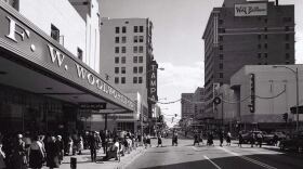 A vintage black/white photo showing F.W. Woolworth to the left with a street in the middle and tall buildings in the background