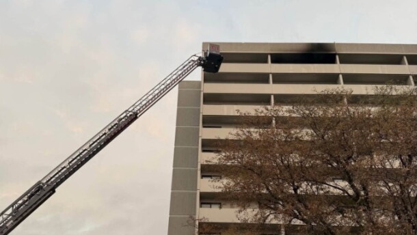 Fire trucks with extended ladder respond to a fire on the upper floor of a multi-story building, with black smoke visible from the windows. Trees and emergency lights are also visible in the scene.