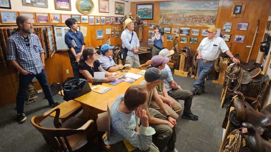 A group of archaeologists, students and community historians sit and stand inside a wood-paneled museum room filled with framed photographs, saddles and ranching artifacts while listening to a presentation about Grant County history.