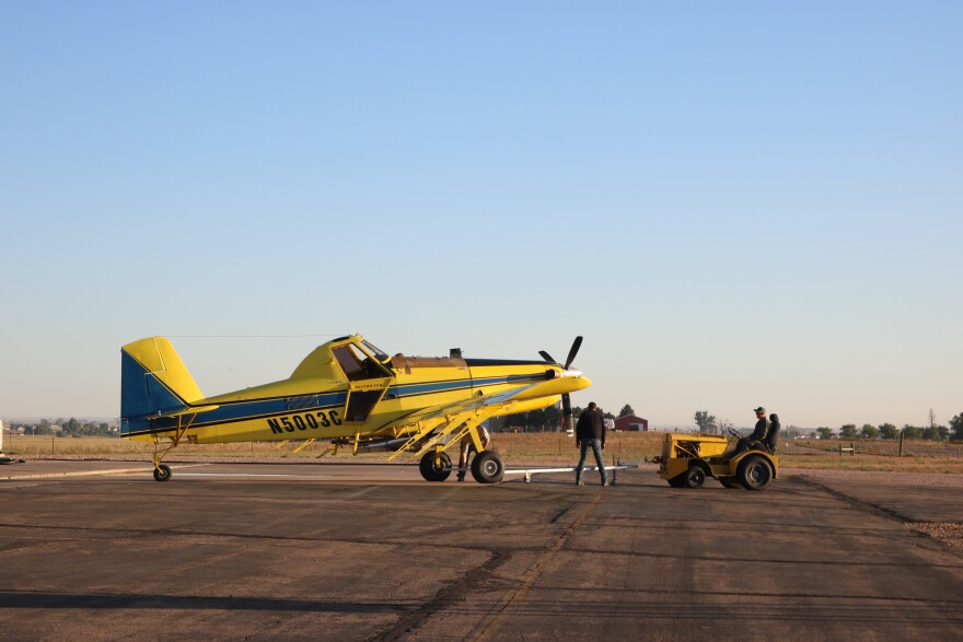 A yellow plane with blue and black stripes sits on a tarmac. A man is in front of it looking at the propeller. There is another man on a yellow tractor that's attached to the plane.