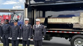 Four firefighters in navy blue uniforms stand at attention near a truck with a side panel open that shows a horizontal beam from the World Trade Center