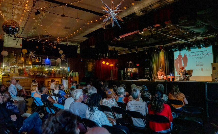 A large audience sits inside a music venue watching two speakers onstage beneath string lights and hanging decorations.