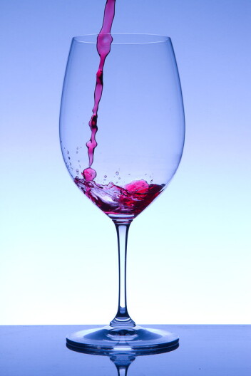 A tall wine glass sits in front of a blue background while a red liquid is poured into it.