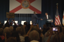 Florida Gov. Ron DeSantis shakes hands with U.S. Rep. Byron Donalds onstage in front of a large Florida flag as audience members watch and take photos.
