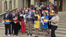 A large group of approximately 25 people pose on the steps of the Michigan State Capitol building on a sunny day. Most hold printed Progress Pride flags — the rainbow flag design incorporating additional chevrons in black, brown, pink, light blue, and white representing LGBTQ+ people of color and transgender individuals. Several people in the front row also hold what appear to be documents or proclamations with black folders. The group is diverse in age and race, dressed in business and business-casual attire. A string of small rainbow flags connects participants across the front row.