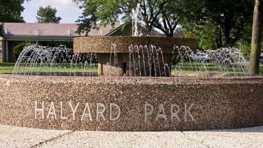 A stone fountain in the middle of a park that says "HALYARD PARK" on the side with a grassy field and one-story building in the background.