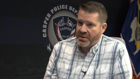 A man wearing a button up plaid dress shirt speaks in front of the Casper Police Department's seal and a Wyoming flag.