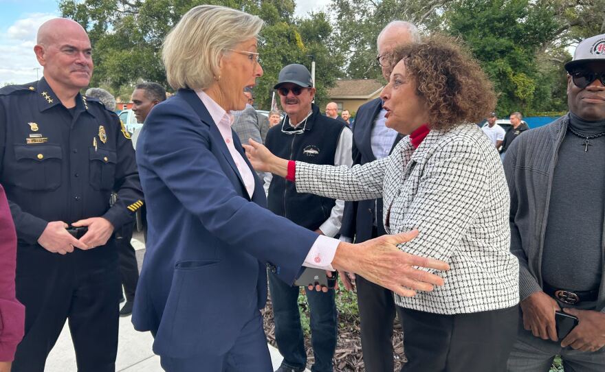 A woman in a navy suit excitedly embracing a woman in a white and black suit while people stand behind them.